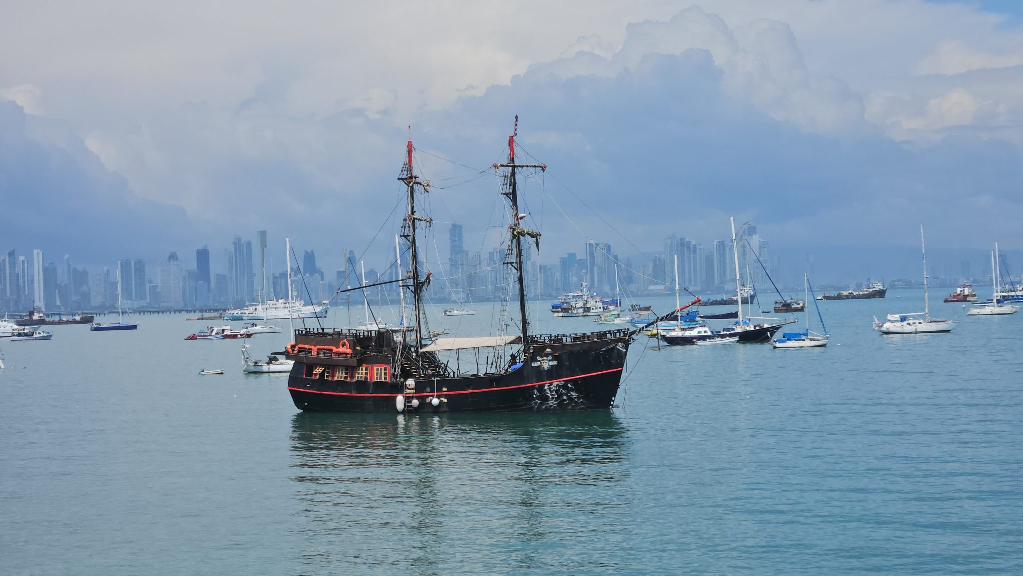 A black pirate ship with red trim and two masts sails in a harbor, surrounded by various boats, with a city skyline of skyscrapers under a cloudy sky.