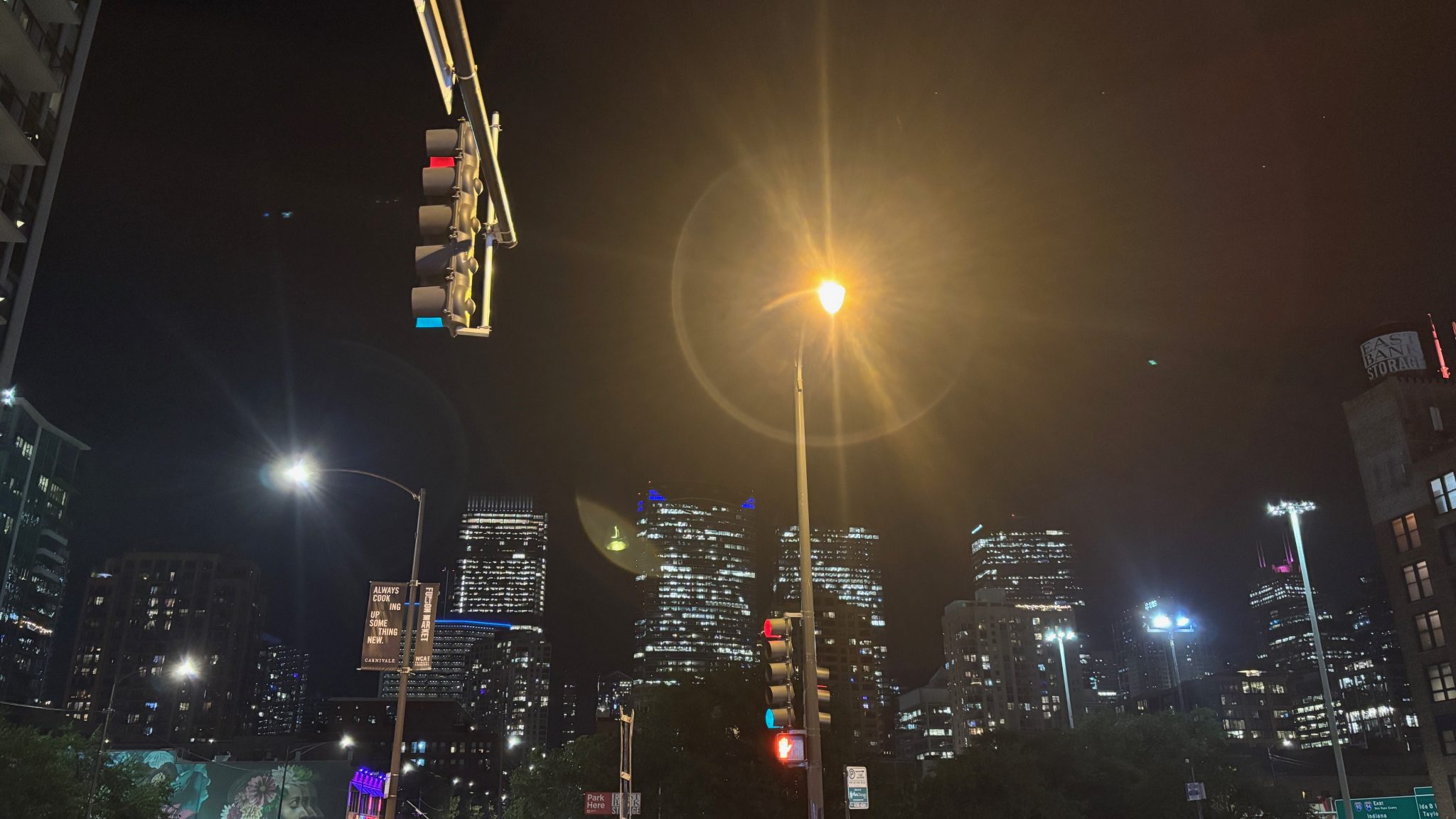 A city skyline at night. In the top left, quite close, is a stop light pointed to the left. In the center is a bright street light with strong lens flare around it.