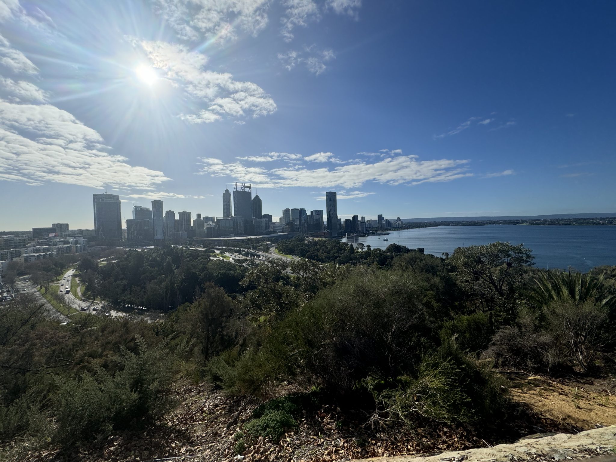 A city skyline viewed from a hilltop, with a bright sun shining in a partly cloudy sky. The city features several high-rise buildings and is situated near a large body of water. Trees and natural vege...