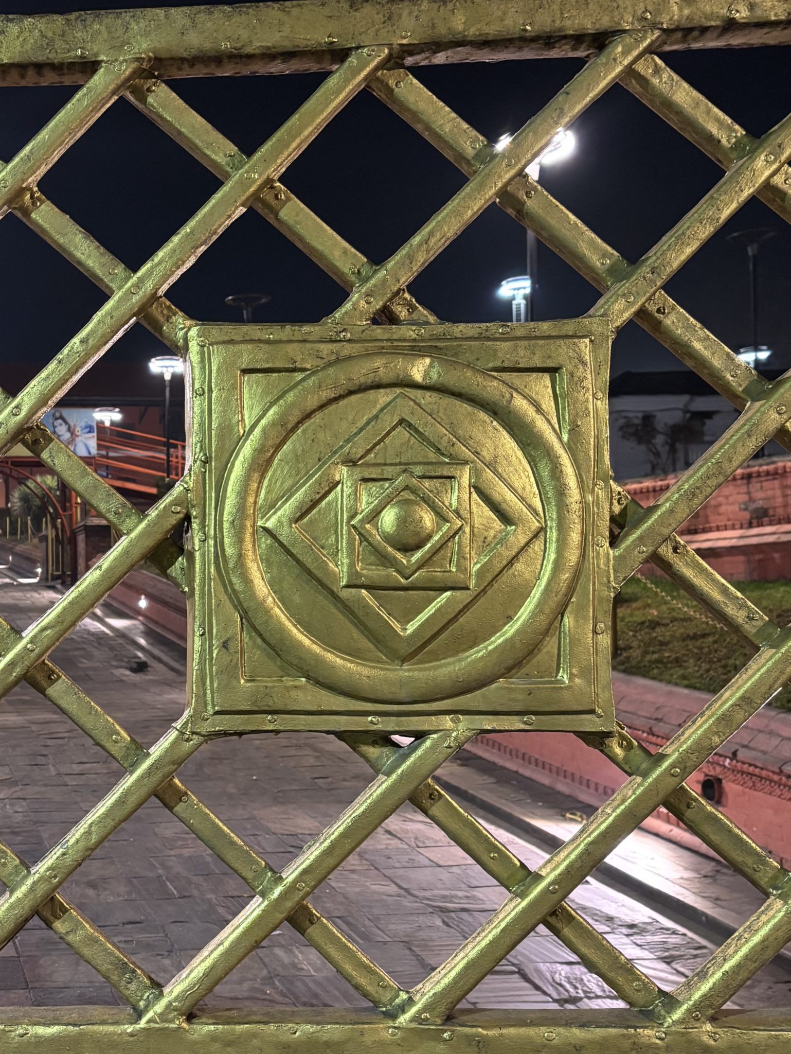 A close-up view of a decorative golden grill with a diamond-patterned design and a central emblem featuring geometric shapes, set against a dark background with dim lighting