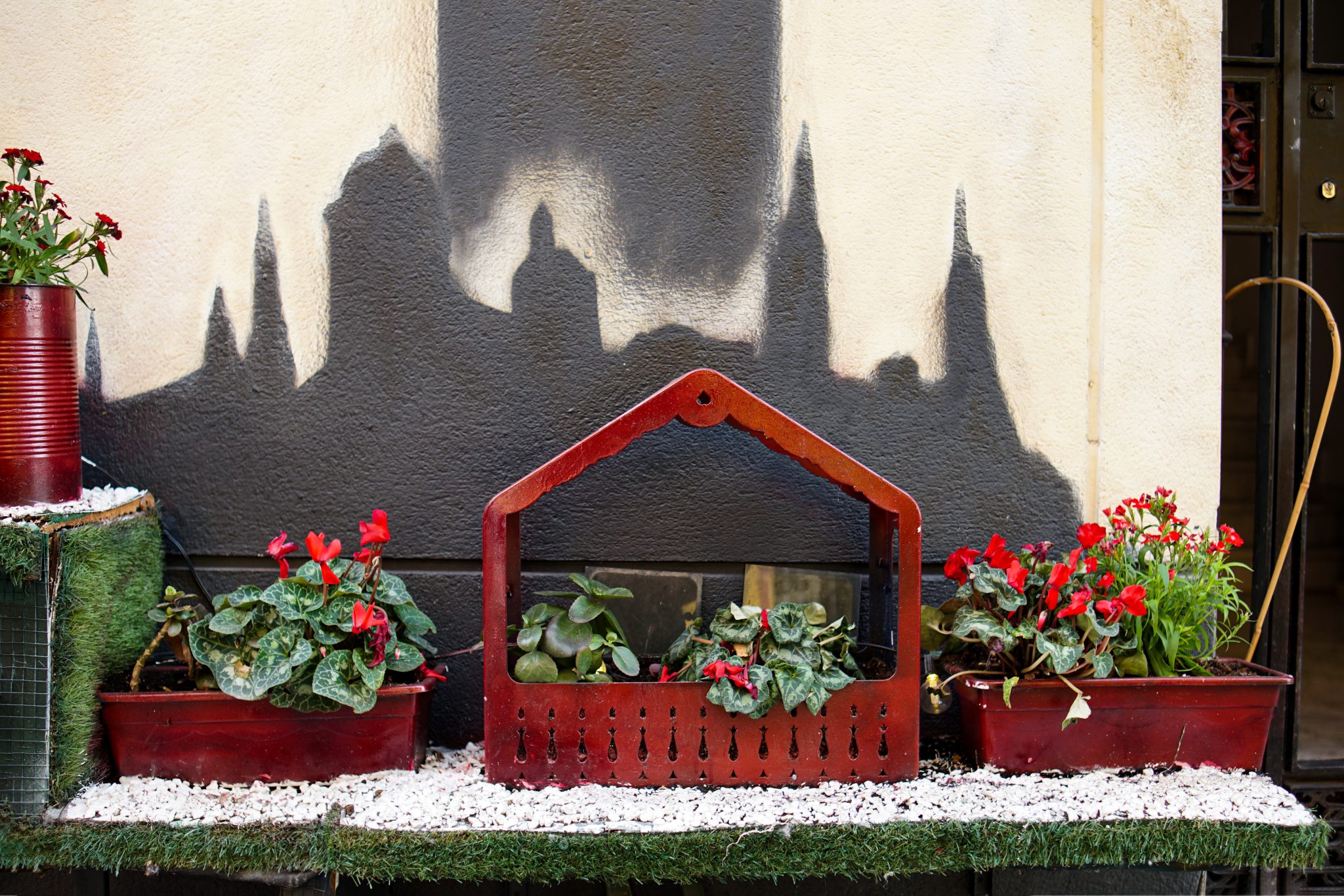 A display of red flowers in pots set against a wall with a mural depicting the silhouette of a city’s skyline. The flowers are arranged around a decorative red wooden frame.