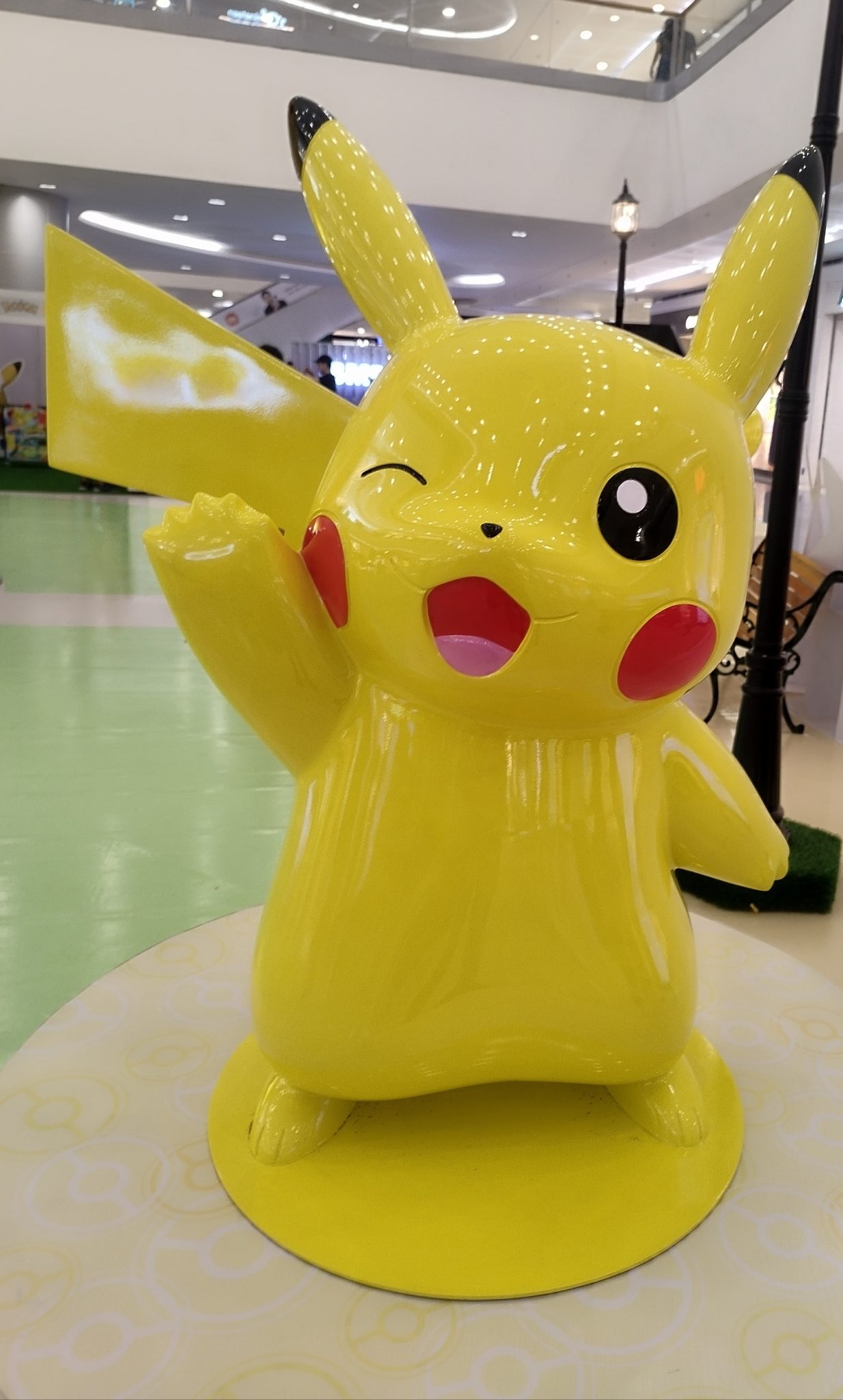A large, cheerful Pokeeee of a yellow cartoon character with long ears, red cheeks, and a lightning bolt-shaped tail, standing on a pedestal in a shopping mall setting.