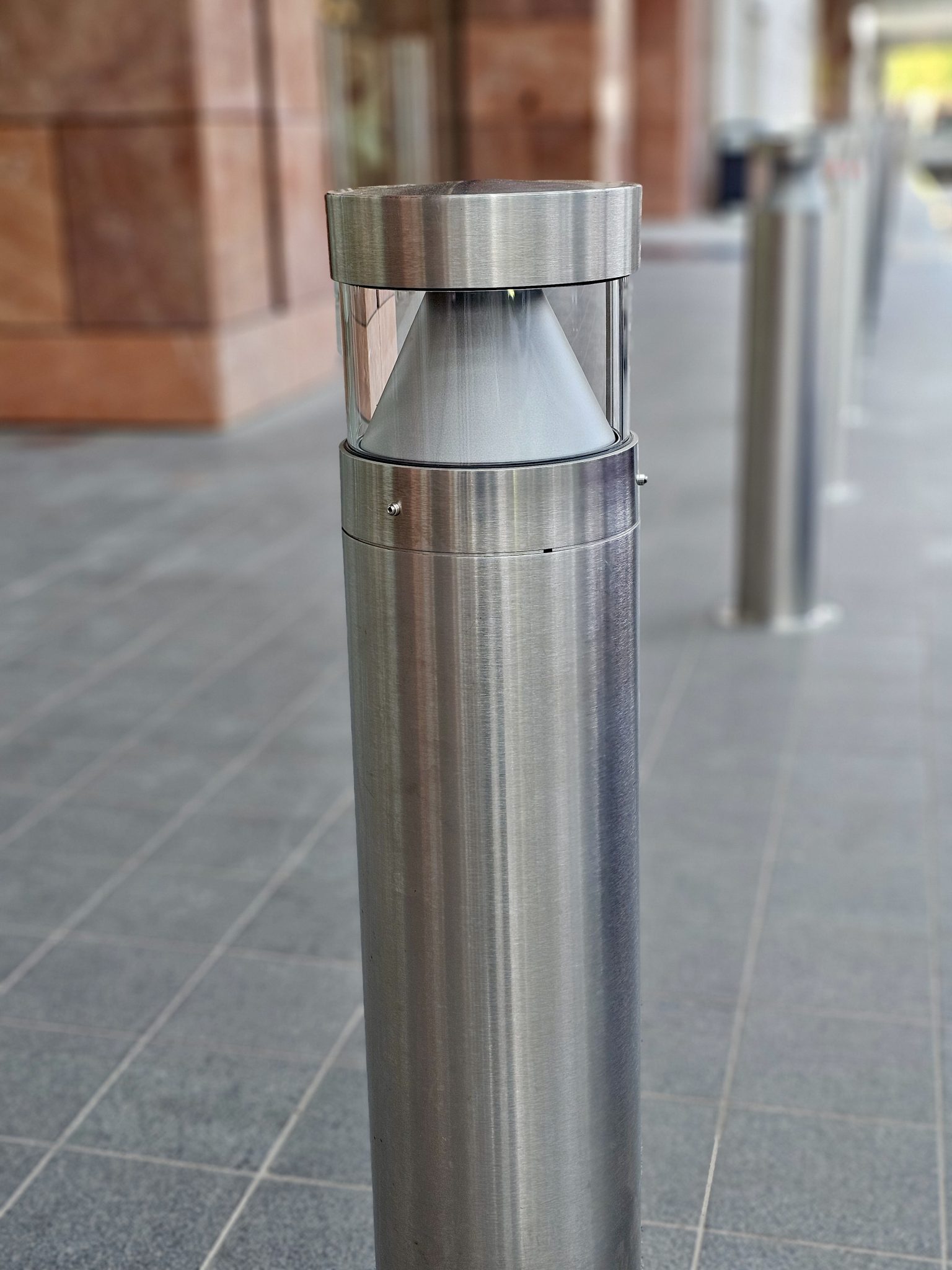 A modern stainless-steel outdoor lamp standing on a tiled walkway in BKC, Mumbai. It is a clean, minimal design that looks sleek and stylish.