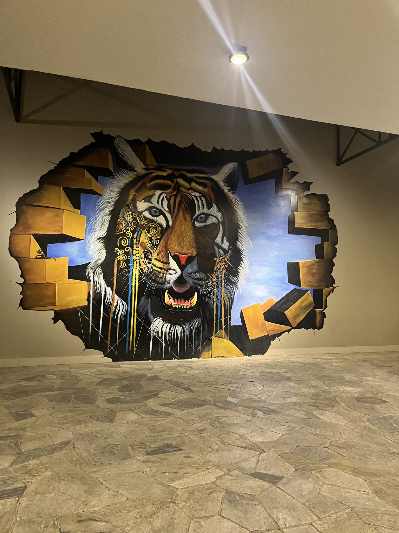 A mural painting of a tiger’s face on a wall, giving the illusion that it is breaking through the surface. The tiger features detailed eyes and prominent teeth, and one side is adorned with intricate...