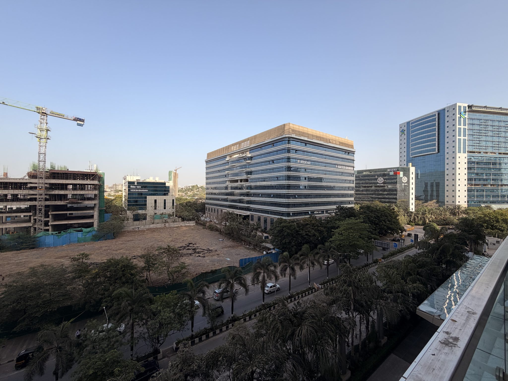 A panoramic view of a modern city skyline with glass office buildings, palm-lined streets, and ongoing construction. The scene captures urban development and a clear blue sky.