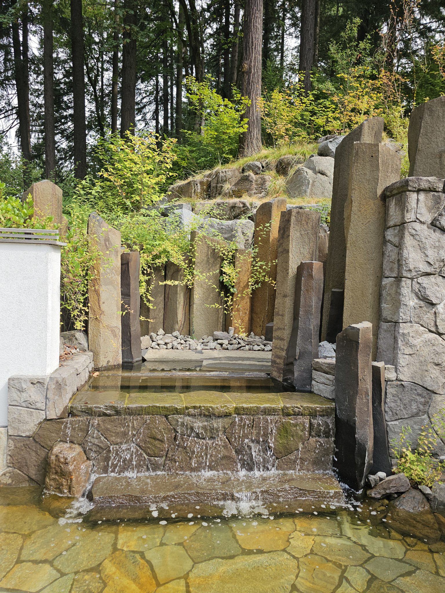 A peaceful stone waterfall with tall basalt columns and forest trees behind it. Captured on a clear day at the Portland Japanese Garden.