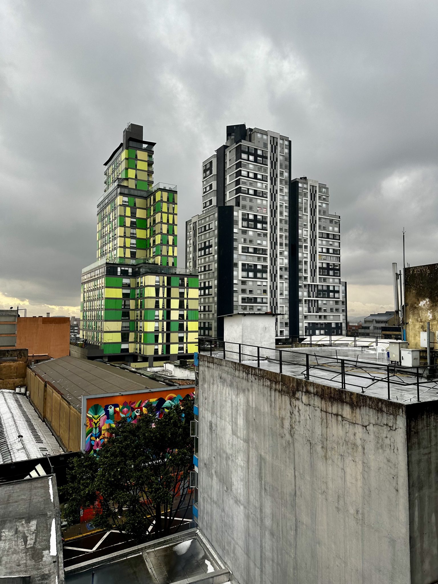 A rooftop view over central Bogotá, Colombia, under a heavy overcast sky. A striking residential tower with green and yellow mosaic panels dominates the left, flanked by grey concrete high-rises. A vi...