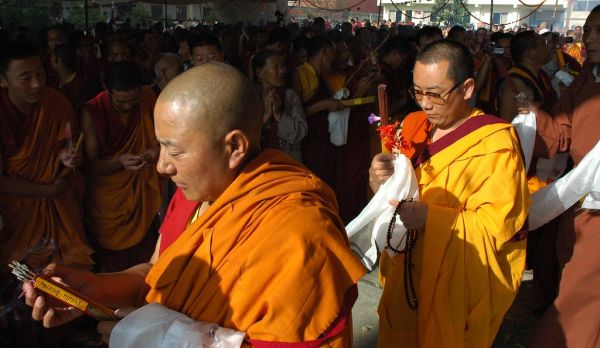 A senior lama wearing Sanghati - bright orange patchwork robes - pulls more incense from the carton, in the Lamdre Bodhisattva Day procession, a monk carries flower, incense, katag and mala, offerings...
