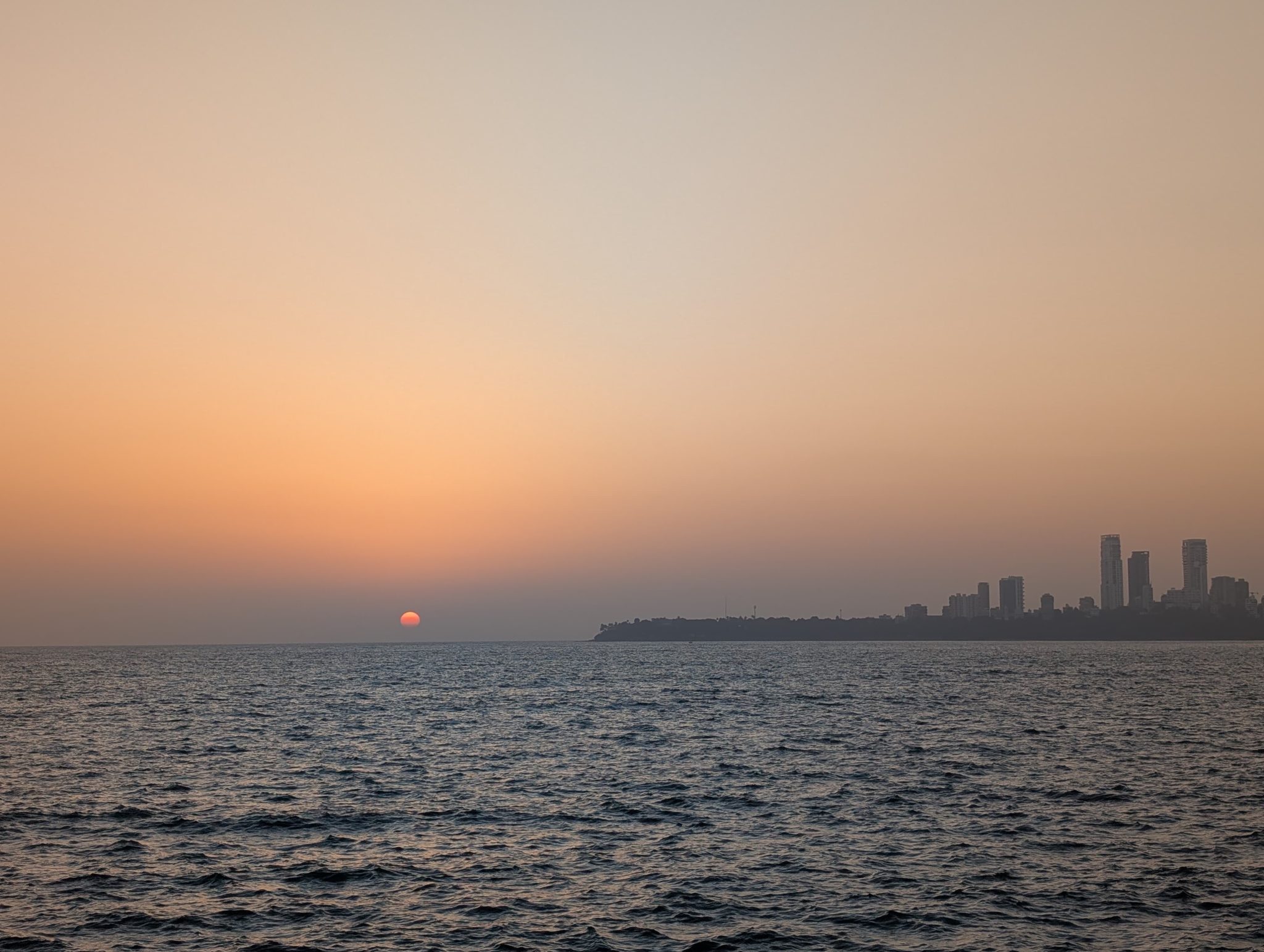 A serene sunset over the ocean, with the sun partially dipped below the horizon, casting a warm glow in shades of orange and pink. The calm waters reflect the colors of the sky, while a city skyline w...