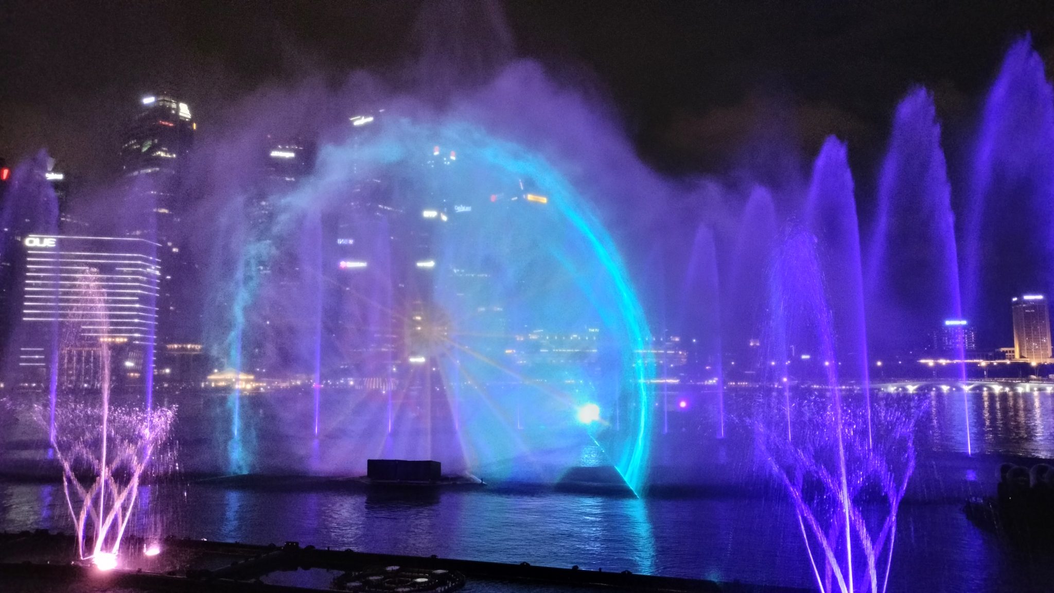 A stunning night view of the Marina Bay Sands Light and Water Show in Singapore, featuring vibrant water fountains illuminated in shades of purple and blue. The mesmerizing water projections create a...