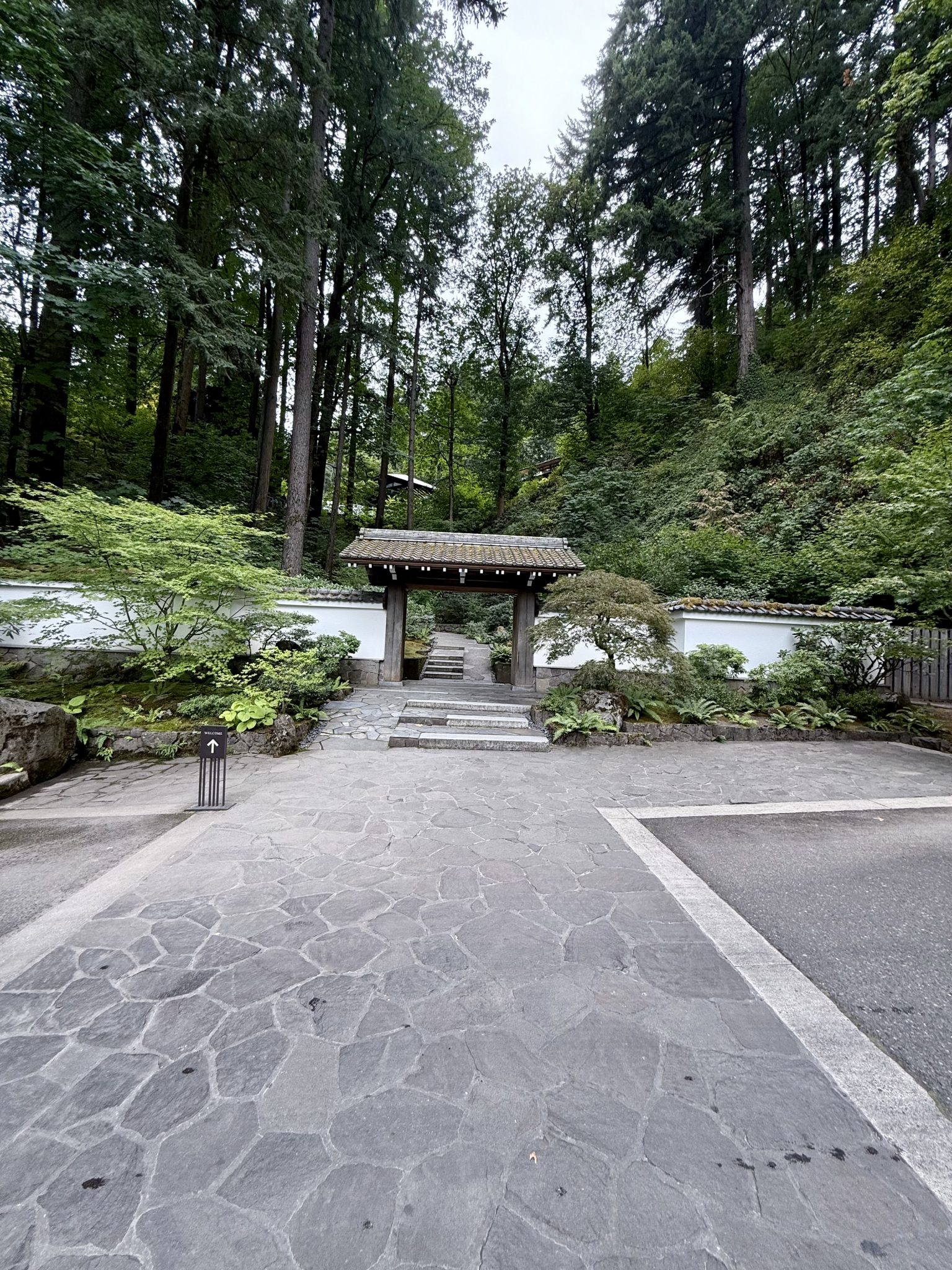 A traditional wooden Japanese gate stands at the entrance of the Japanese Garden, Portland, framed by stone paths and tall forest trees in the background.