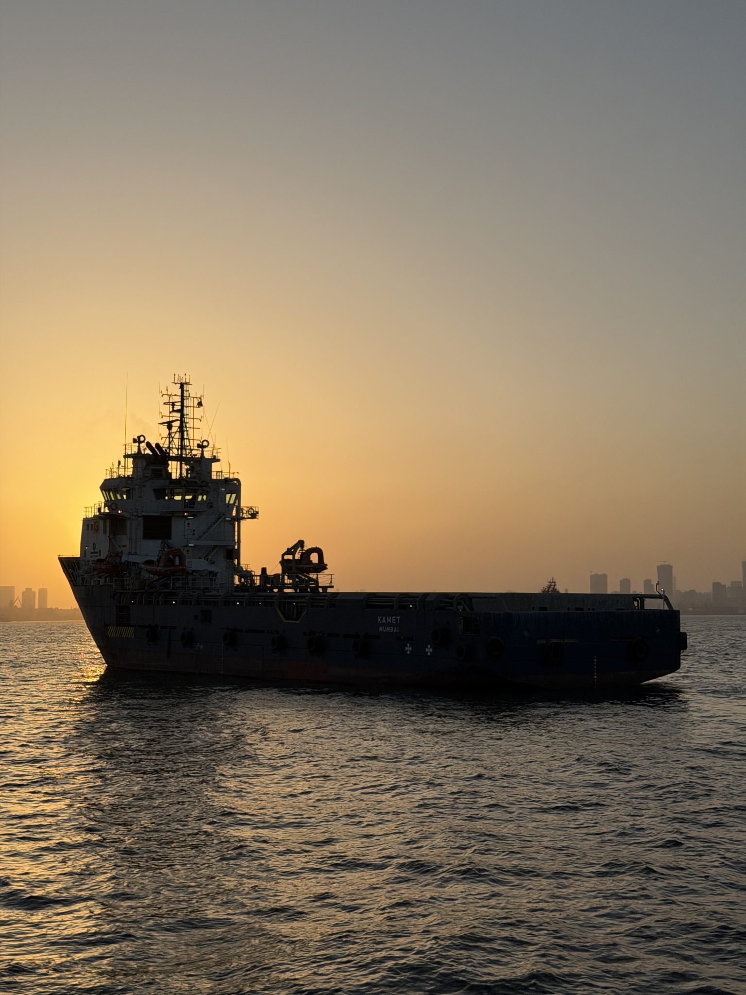 A tugboat/supply ship in harbor off the coast of India. Itβs silhouetted against the setting sun, and you can see a city skyline in the distance.