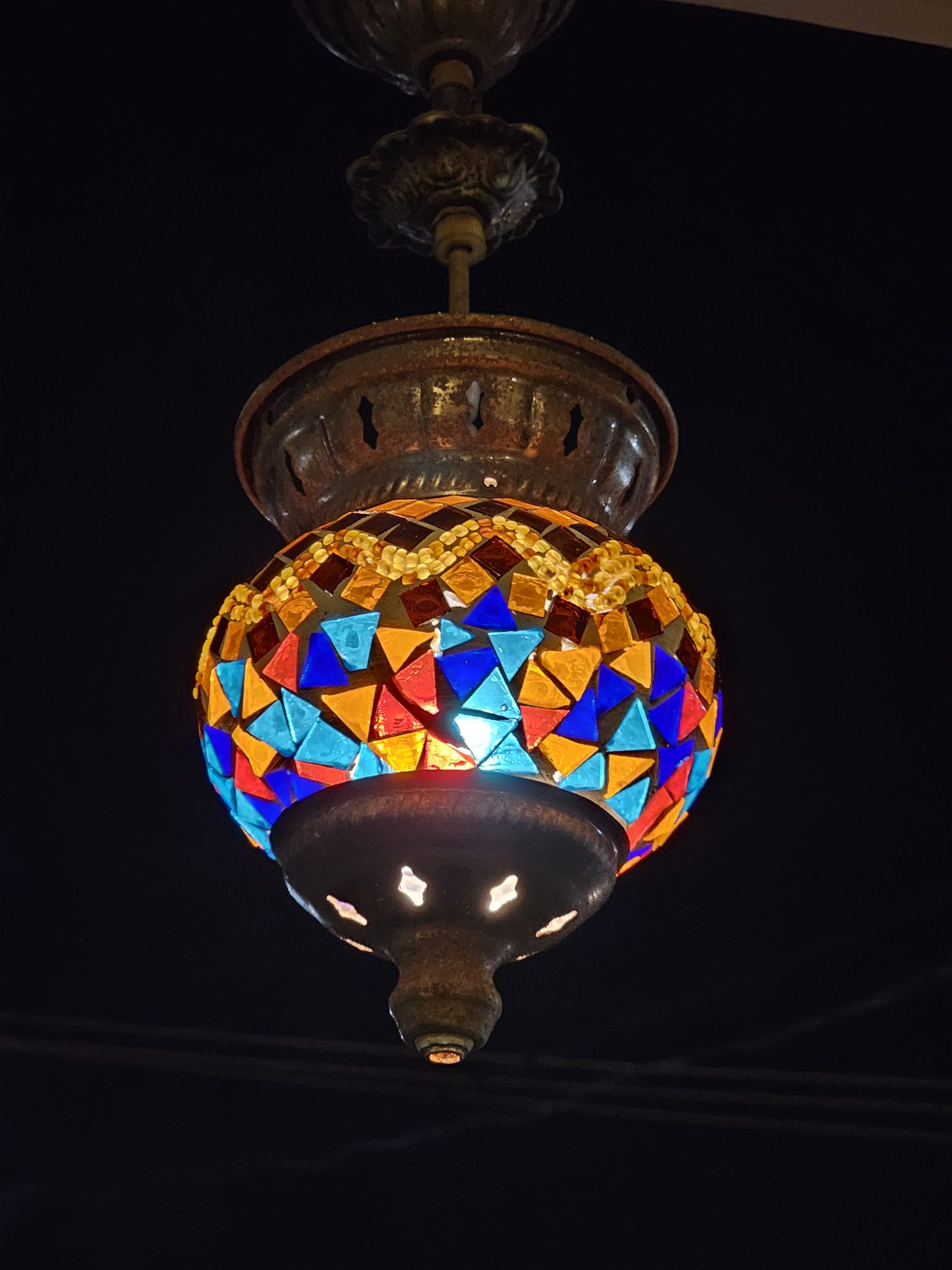 A Turkish mosaic lamp hanging from a ceiling fixture against a dark background. The lamp features a spherical lampshade adorned with colorful, geometrically shaped glass pieces, and it emits a warm gl...