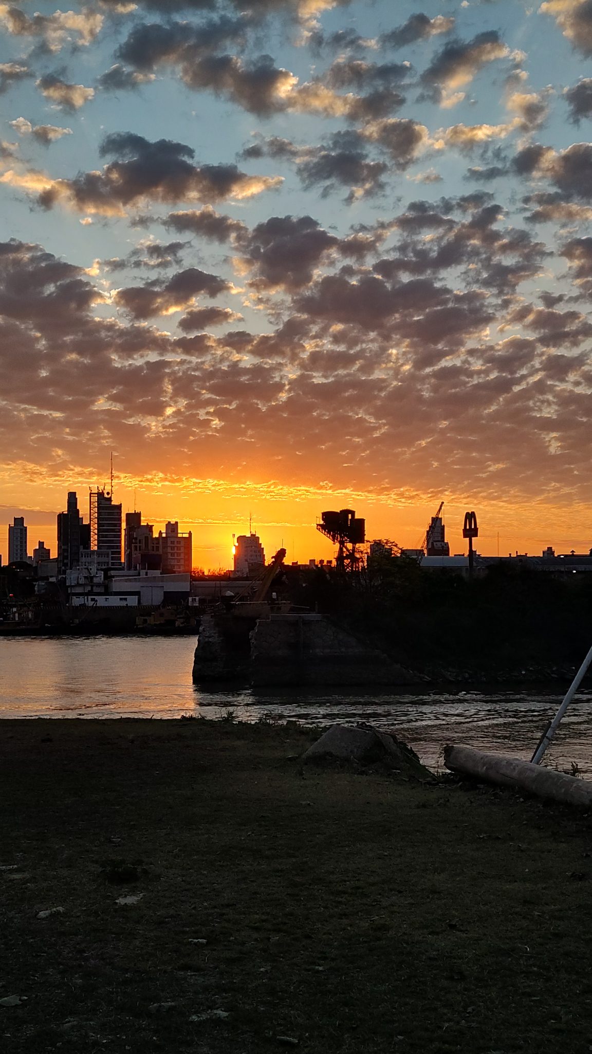 A vibrant sunset illuminates the sky with hues of orange and yellow, casting a silhouette over a city skyline near a body of water. Scattered clouds are tinged with the fading sunlight, and there is a...