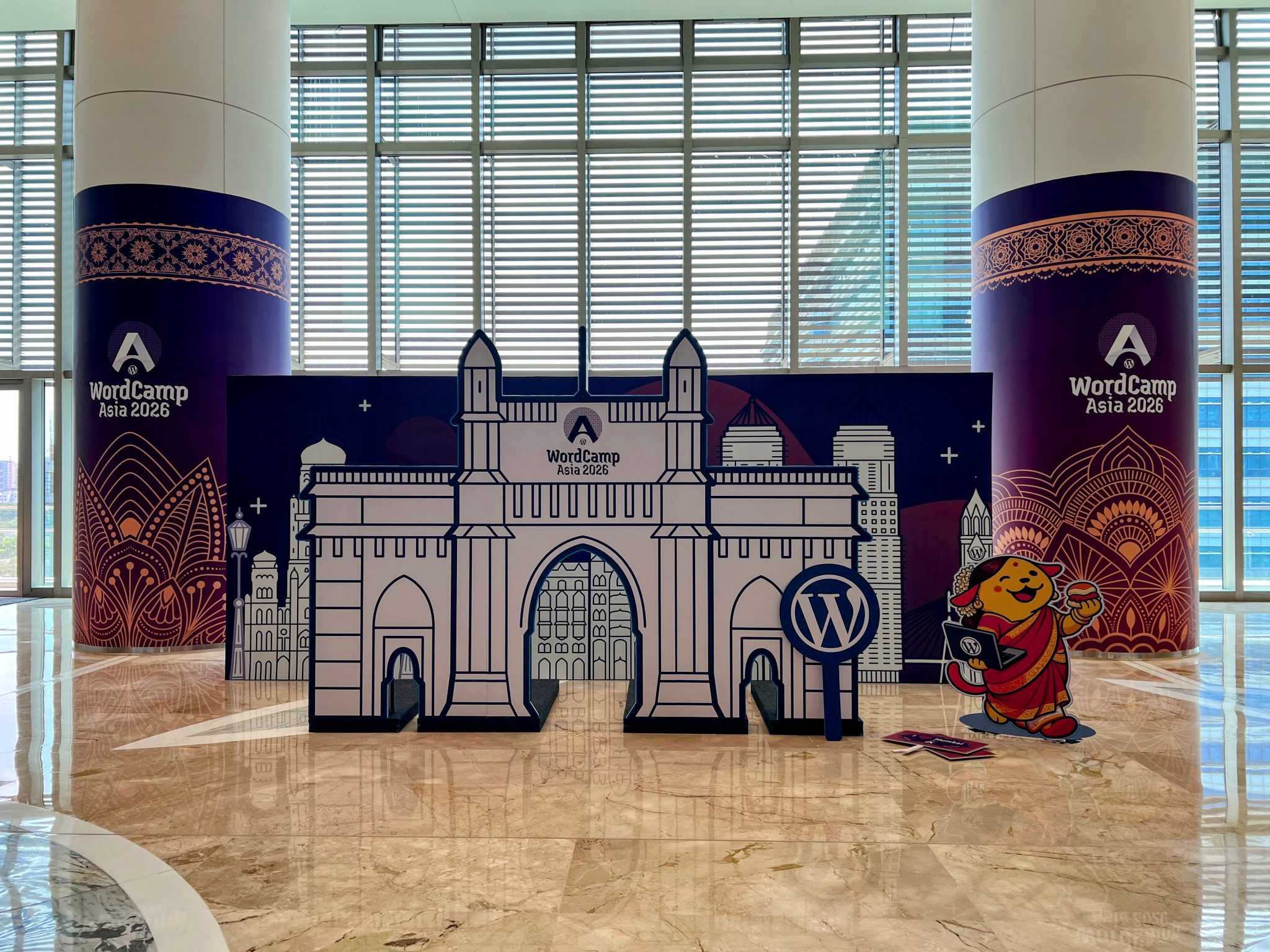 An event photo backdrop featuring a large cutout of the Gateway of India, a WordPress logo sign, and a cartoon character illustration, set between two purple “WordCamp Asia 2026” pillars.