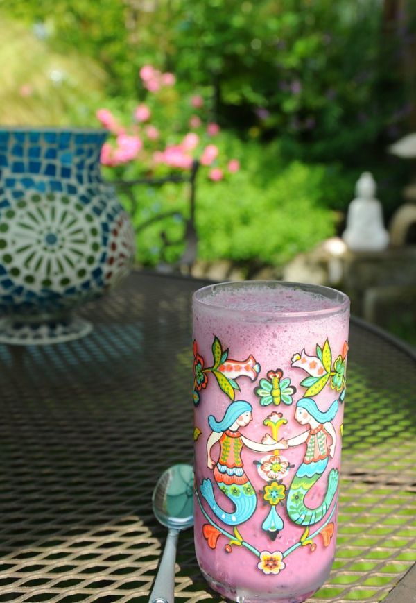 Boysenberry milkshake in a tumbler decorated with two mermaids (World Market), spoon, mesh patio table, blue glass candle lamp, white Buddha, Japanese lantern, pink carpet roses, A Garden for the Budd...