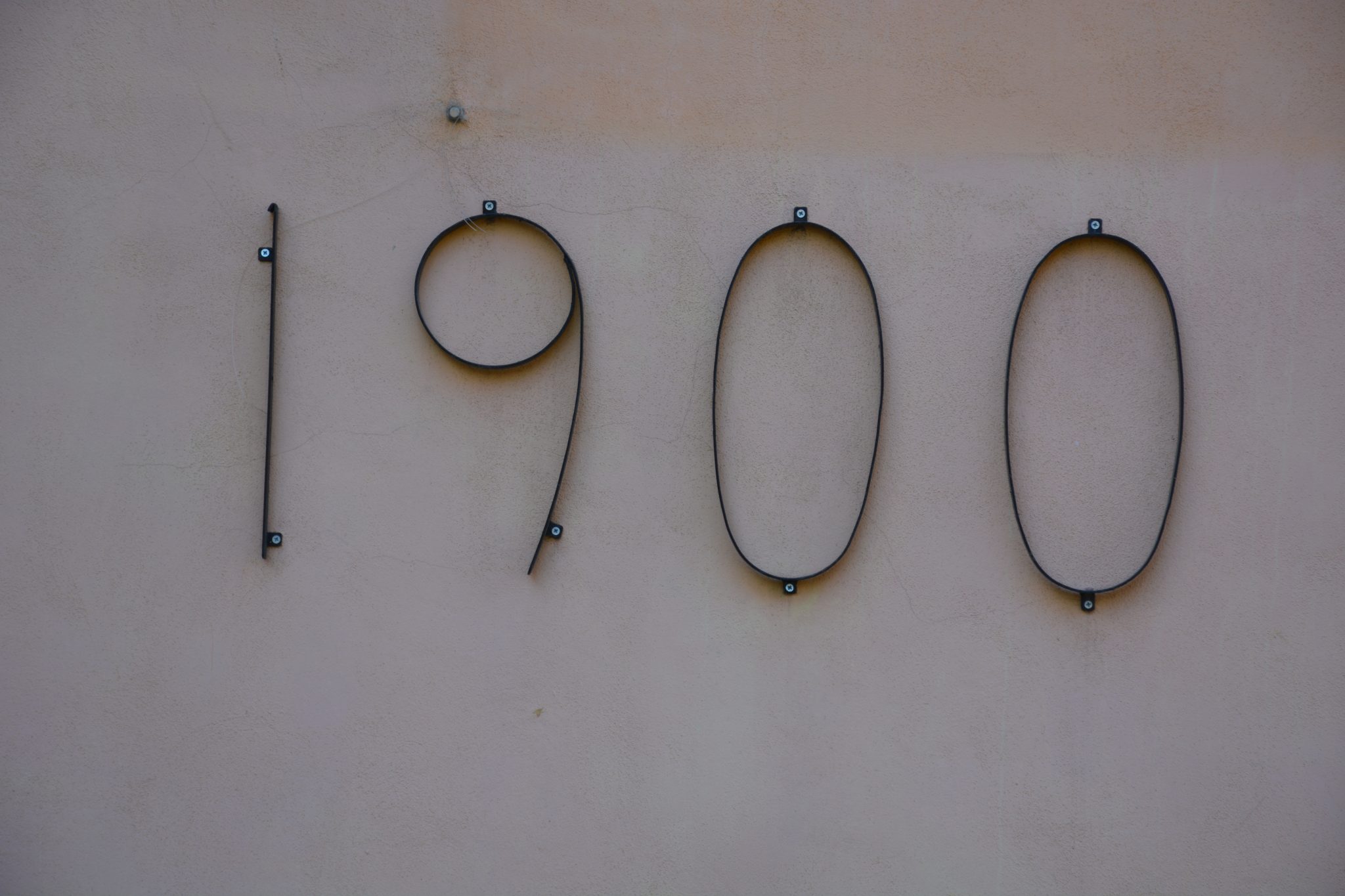 Elegant wrought iron numbers ‘1900’ mounted on a pastel-colored textured wall, likely indicating the building’s construction year. Captured in Cyprus, the minimal design evokes a quiet sense of histor...