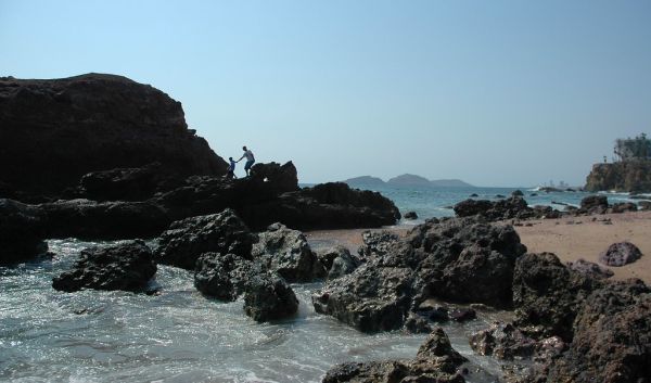 Father and son holding hands for balance while walking on black beach rocks, summer, clear blue sky, Pacific shore, South Mazatlan, Sinaloa, Mexico