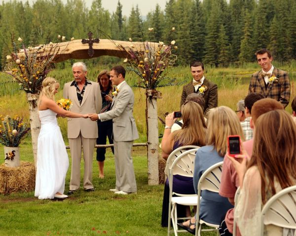Jessie about to marry her beau, Chris, officiant, groomsmen in vintage clothing, photographer, audience with camera phones, country style, cross topping the white birch wedding arch, Fairbanks, Alaska...