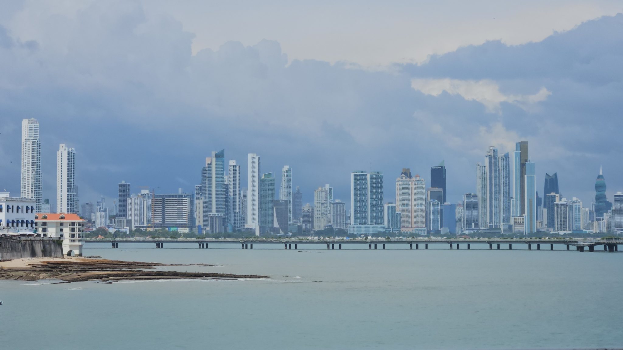 Panama City skyline with numerous high-rise buildings overlooking a calm body of water, featuring a long pier and a cloudy sky, viewed from a waterfront location.
