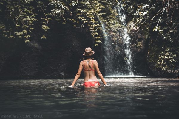 Rear view of Young woman tourist with straw hat in the deep jungle with waterfall. Real adventure concept. Bali island.