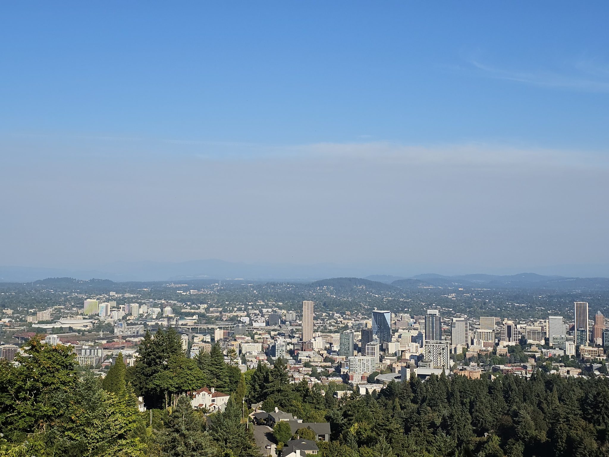 Scenic view of downtown Portland city skyline surrounded by trees and distant hills, taken from the Pittock Mansion in the evening.