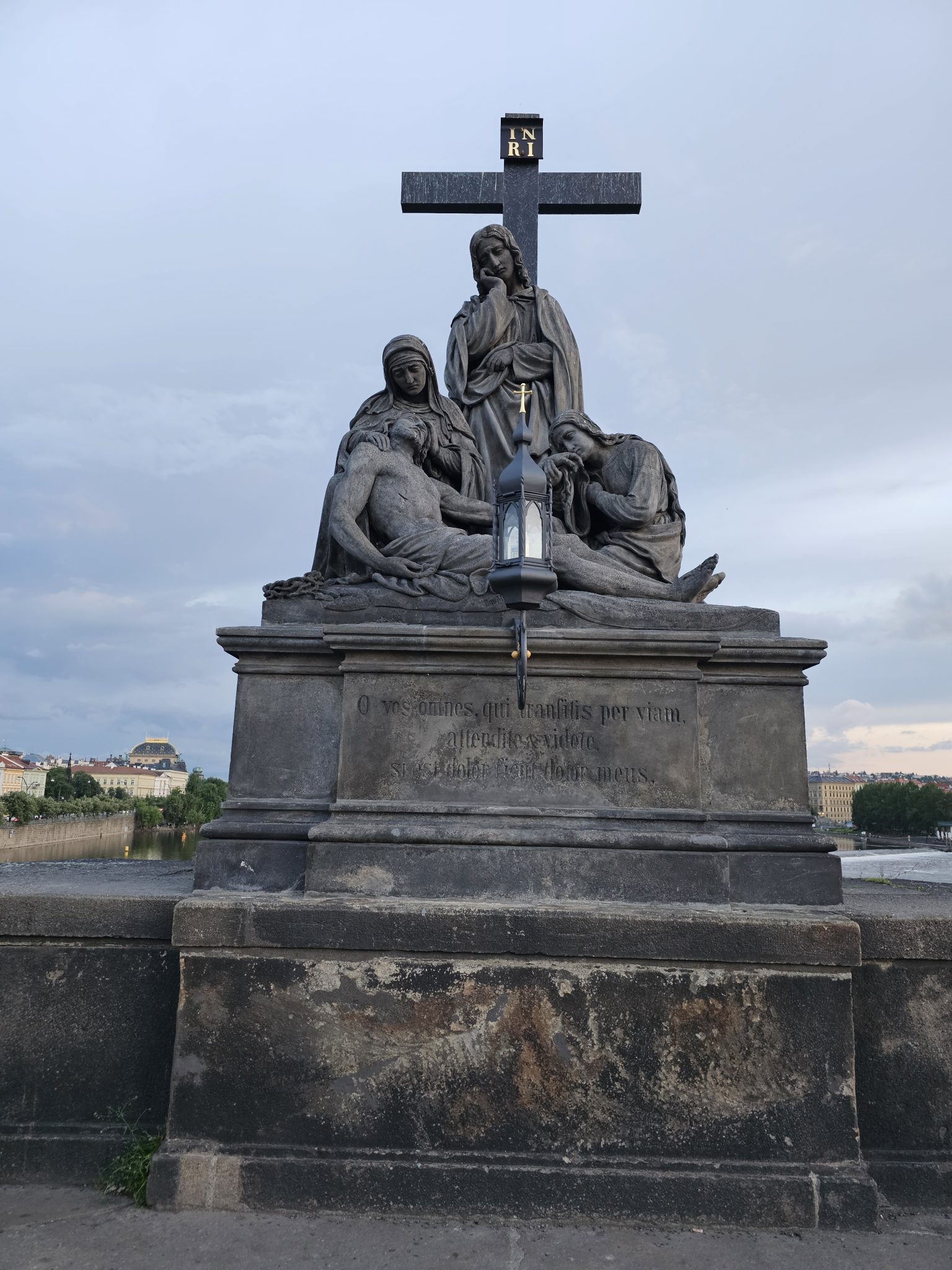 The statue of Pietà on the Charles Bridge in Prague depicts the Virgin Mary cradling the deceased Jesus Christ, with other figures present. The statue is made of dark stone and stands on a pedestal wi...