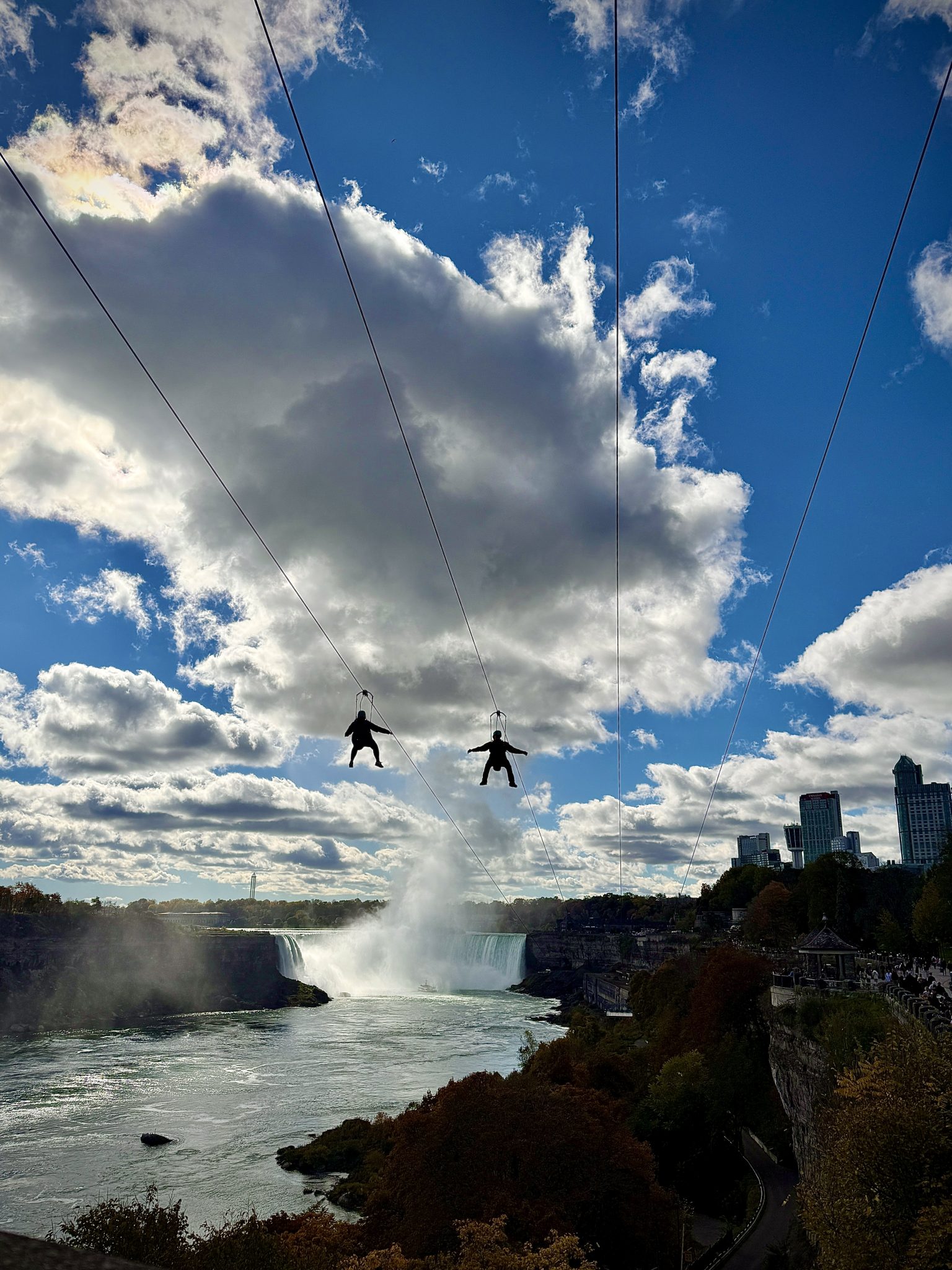 Two people ziplining high above Niagara Falls on parallel cables, silhouetted against a bright blue sky with dramatic clouds. Below them, mist rises from the powerful waterfall, while the city skyline...