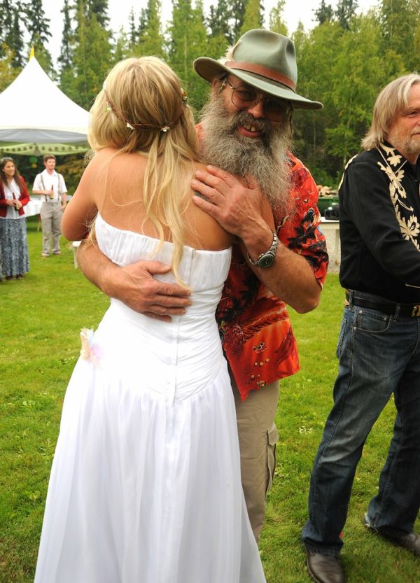 Well wishes! Jessie in her white wedding dress hugs Dave Klumb, renown maker of dog sleds, wearing a hat & beard, tent, reception line, Wedding of Jessie and Chris, outside, Fairbanks, Alaska, USA