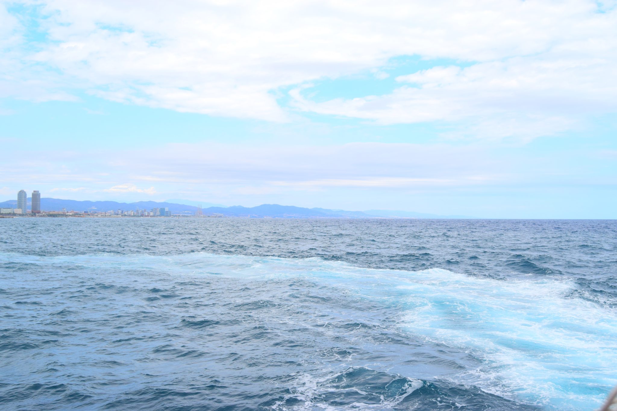 Wide view of the deep blue sea with boat wake foam, featuring a distant coastal city skyline under a cloudy sky.