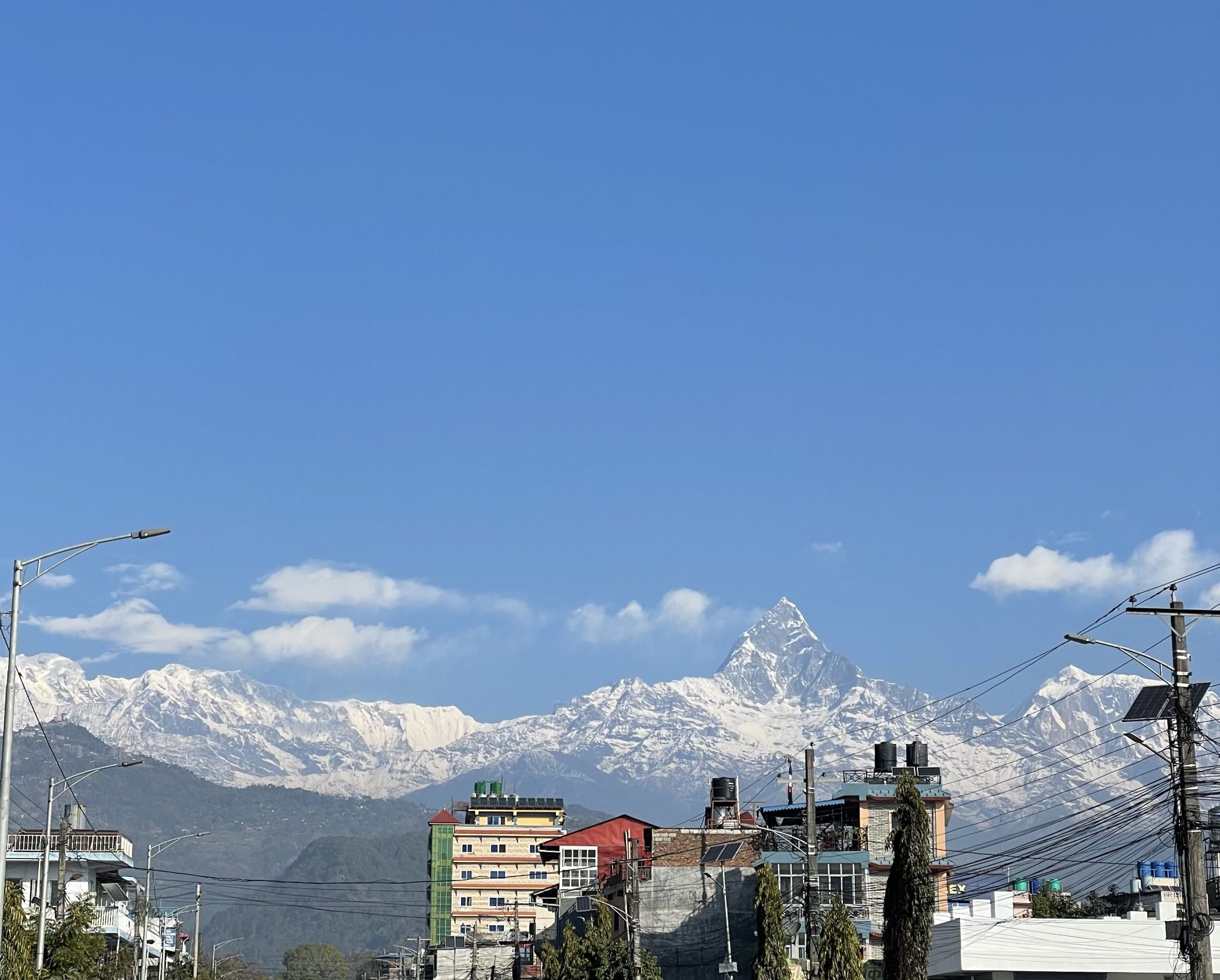 A bright daytime view of a city skyline featuring buildings of varying heights and intersecting utility poles, set against the backdrop of a massive, majestic snow capped mountain peak under a clear b...