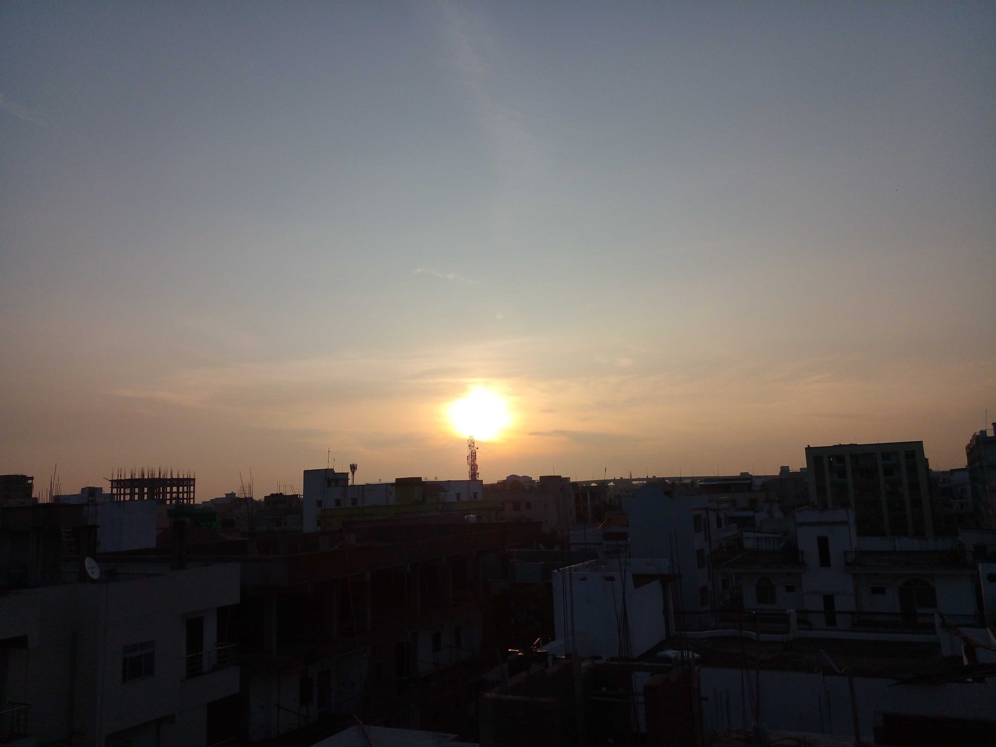 A city skyline at sunset with the sun low in the sky, silhouetting various buildings and structures against a backdrop of a clear sky with subtle orange hues.