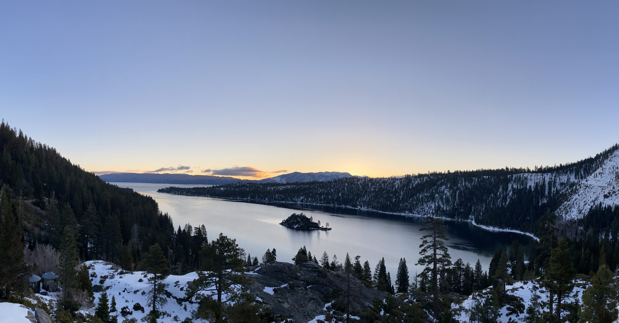A wide panoramic vista of Emerald Bay and Fannette Island at Lake Tahoe during winter. Snow-dusted evergreen forests cover the steep mountain ridges that frame the calm, reflective waters, while the w...
