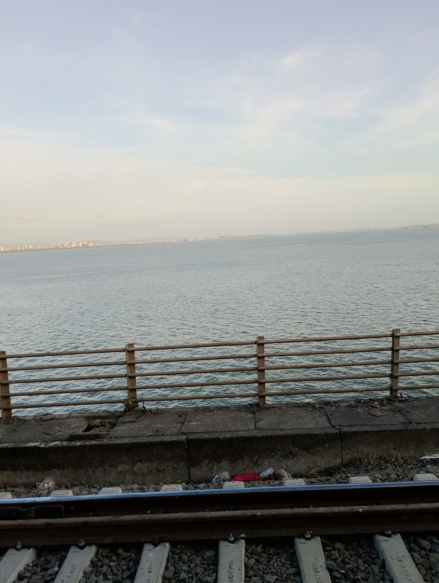 Calm seascape with a distant city skyline under a clear sky. A wooden fence and railway tracks in the foreground add contrast to the serene water view.
