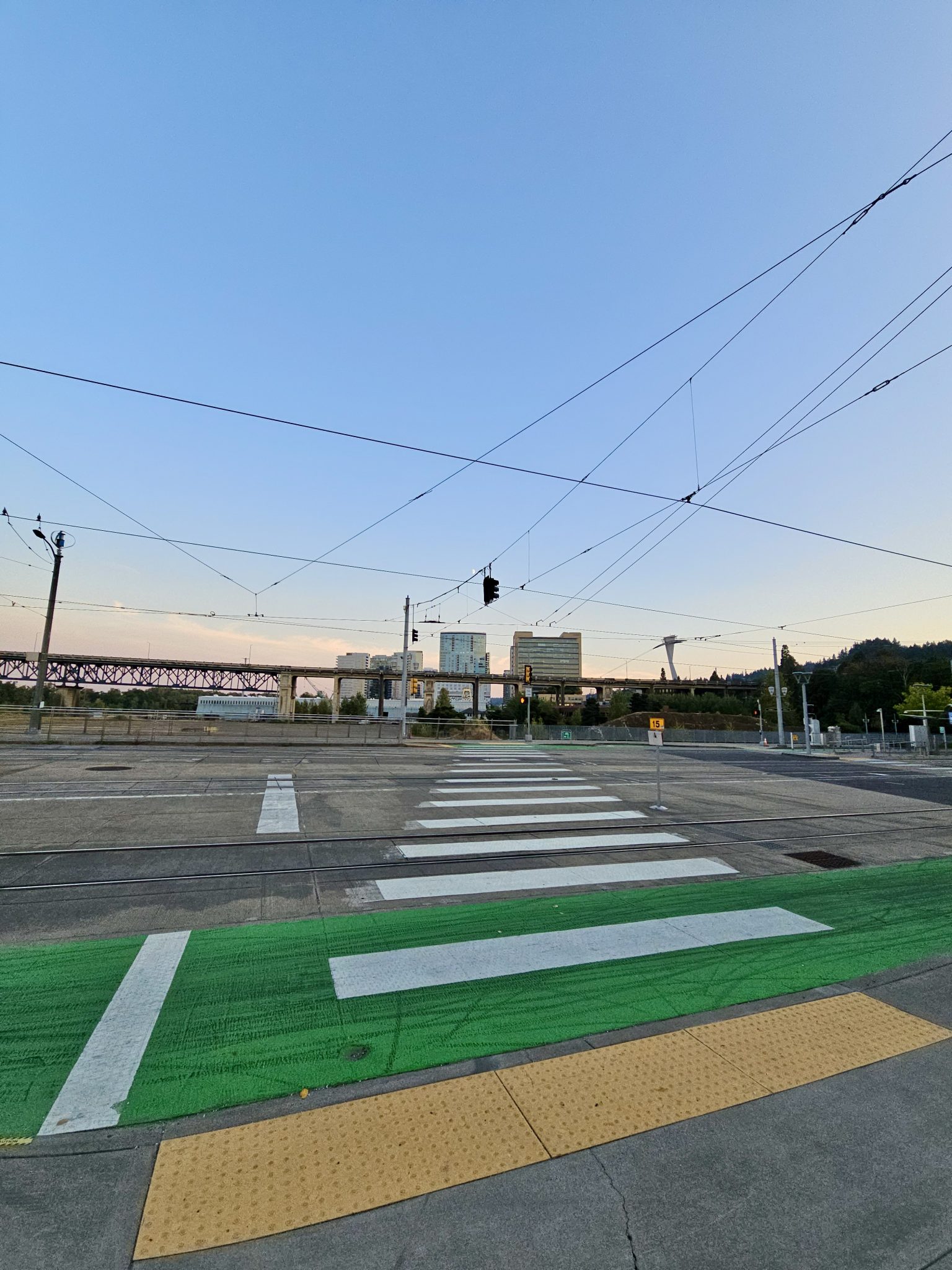 Tram wires above a street intersection near Tilikum Crossing Bridge in Portland. Green bike lane, crosswalk, and city skyline in the distance.